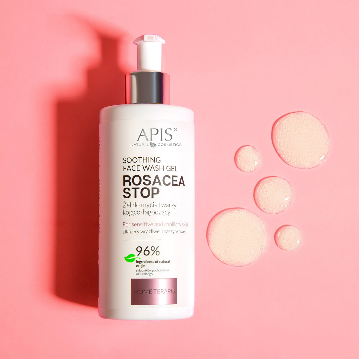 A white pump bottle of APIS Rosacea Stop Soothing Face Wash Gel with a silver collar, set against a solid pink background with decorative droplets of the gel.