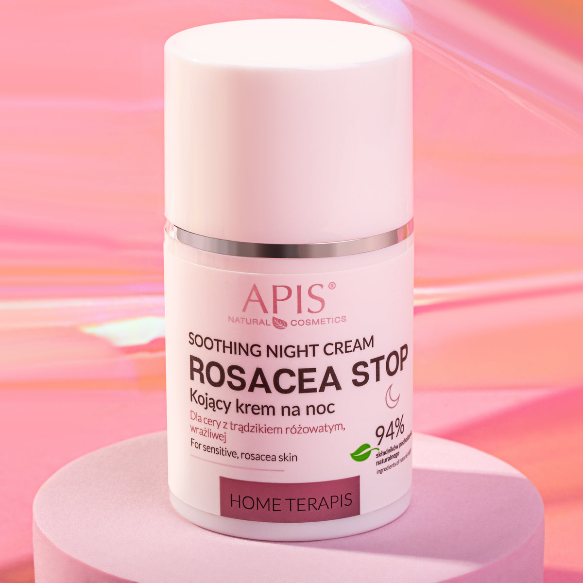 A white cosmetic bottle of APIS Soothing Night Cream Rosacea Stop with a pink cap, displayed on a pink pedestal against a pink background.